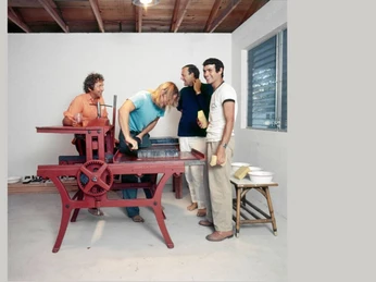 FÜNF FREUNDE In einem hellen Raum versammeln sich vier Personen um eine rote Druckmaschine.In a bright room, four people gather around a red printing press.