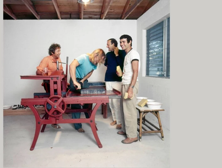 FÜNF FREUNDE In einem hellen Raum versammeln sich vier Personen um eine rote Druckmaschine.In a bright room, four people gather around a red printing press.