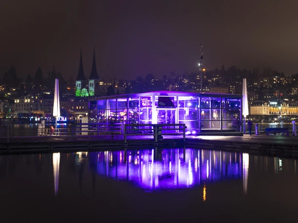 Purple-lit Lilu Village pavilion by Lake Lucerne at night, reflected in the water