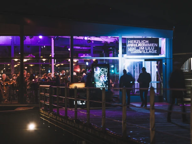 Abendlicher Eingang zum Lilu Village in Luzern, beleuchtet in Pink und Blau mit Gästen