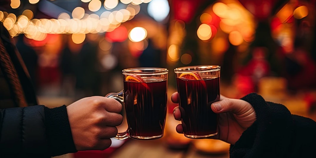 Punschabend zwei Personen, die sich zuprosten mit einer gläsernen Tasse, gefüllt mit Glühwein. Im Hintergrund sieht man Lichterketten und einen Weihnachtsmarkt