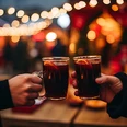 Punschabend zwei Personen, die sich zuprosten mit einer gläsernen Tasse, gefüllt mit Glühwein. Im Hintergrund sieht man Lichterketten und einen Weihnachtsmarkt