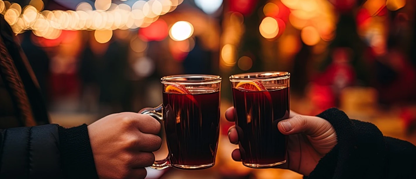 Punschabend zwei Personen, die sich zuprosten mit einer gläsernen Tasse, gefüllt mit Glühwein. Im Hintergrund sieht man Lichterketten und einen Weihnachtsmarkt