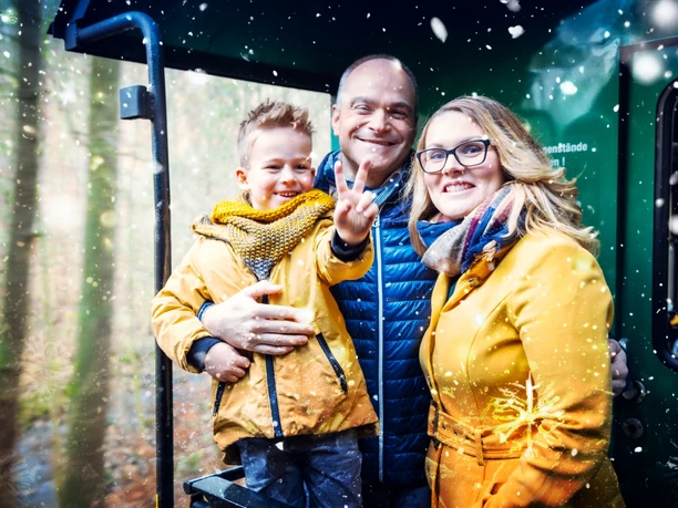 251114_PI_BBLG_Motiv Bimmelbahn und Lichterglanz_Foto Lars Neumann.jpg Familie in gelben Wintermänteln und Schals, lächelnd in einer historischen Bahn, umgeben von Lichtern.