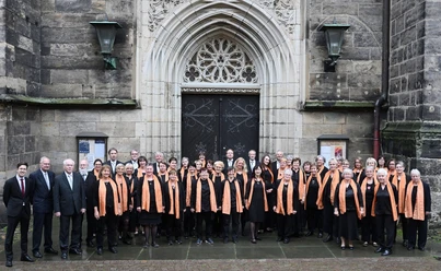 Pirna-Chor_030-1506-24_028Pirna-Chor_030-1506-24.JPG Eine Gruppe von Chormitgliedern steht vor einem prunkvollen, gotischen Kirchenportal.