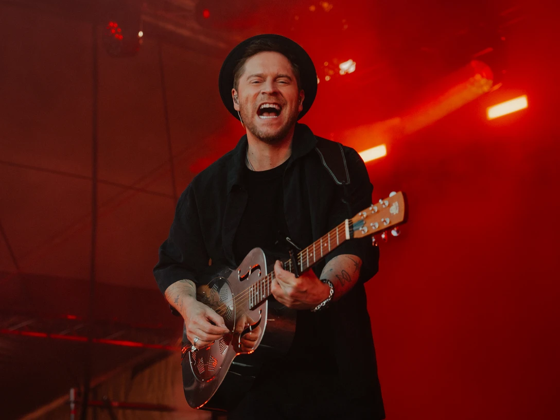 Johannes Oerding in Büsum Ein Mann in schwarzer Kleidung und Hut mit Gitarre auf einer rot beleuchteten Bühne.