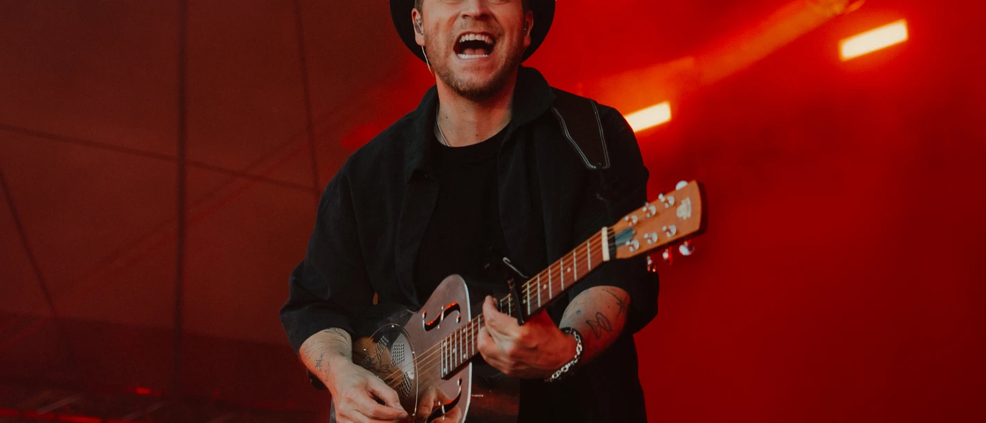 Johannes Oerding in Büsum Ein Mann in schwarzer Kleidung und Hut mit Gitarre auf einer rot beleuchteten Bühne.