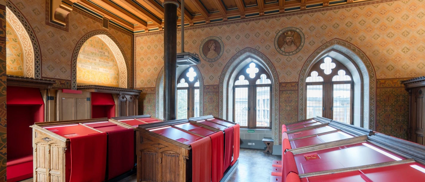 Historischer Raum mit bogenförmigen Fenstern und hölzernen Tresen, die von roten Tüchern überzogen sind.