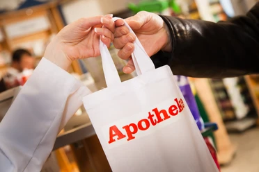 Iemand overhandigt in een apotheek een witte tas met het rode opschrift „Apotheke“.