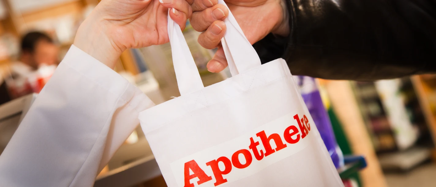 Iemand overhandigt in een apotheek een witte tas met het rode opschrift „Apotheke“.