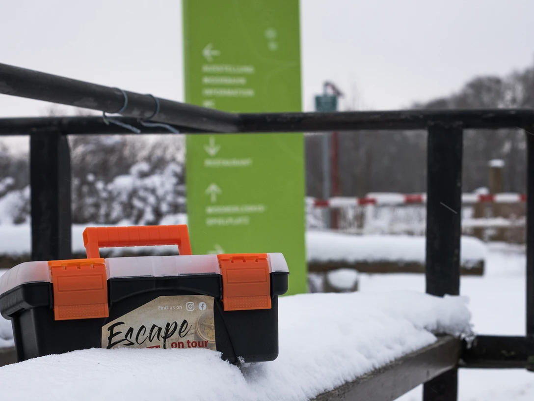 Escapekoffer Ahlenmoor im Schnee Schwarzer Escapekoffer mit orangefarbenem Griff im verschneiten Gelände vor grünem Wegweiser.