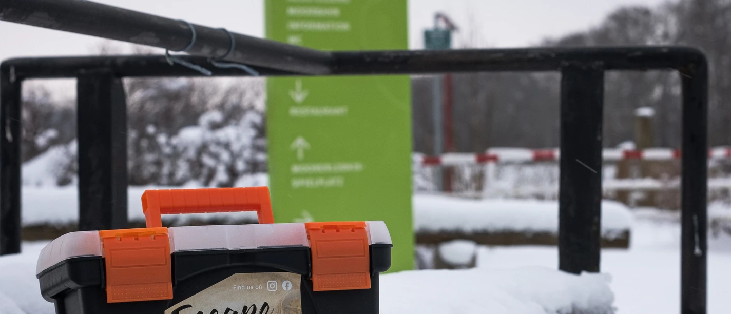 Escapekoffer Ahlenmoor im Schnee Black escape suitcase with orange handle in snowy terrain in front of a green signpost.