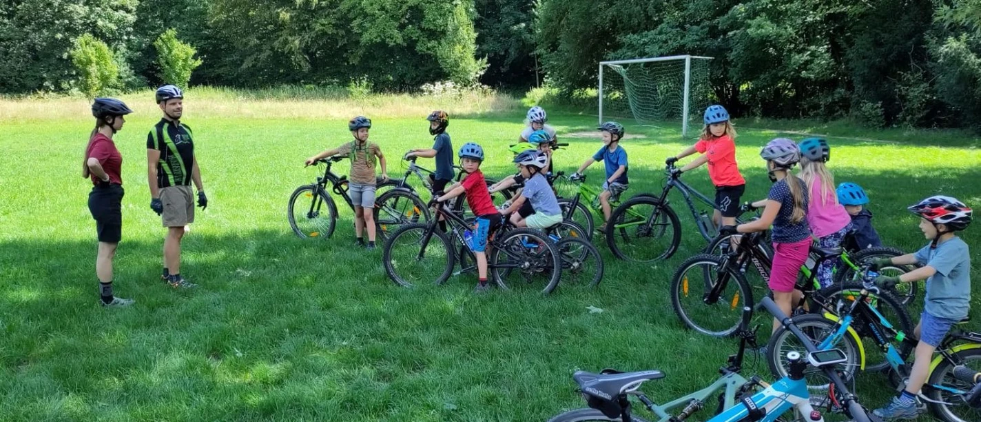 Kinder mit Radhelmen und Rädern auf einer Wiese mit ihren Fahrtrainern