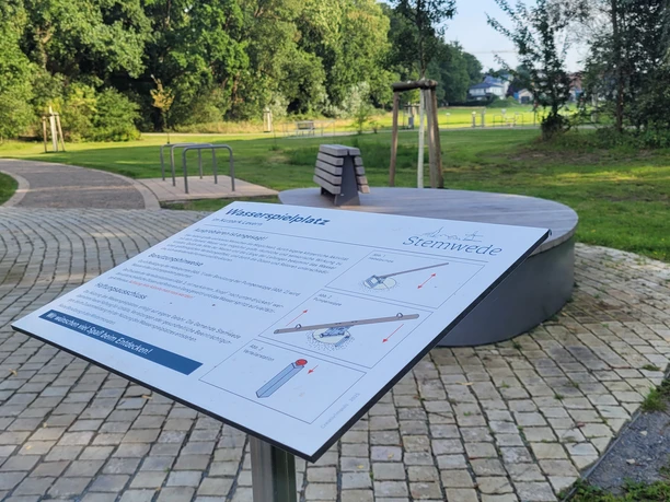 Wasserspielplatz Kurpark Levern Infotafel zum Wasserspielplatz im Kurpark Levern vor rundem Wasserbecken und Grünanlage.