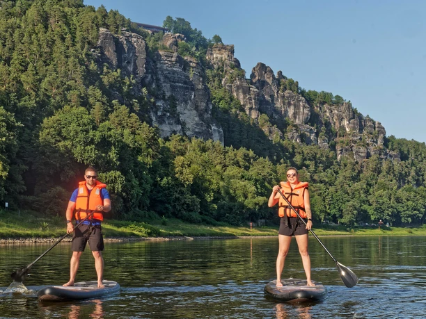 Zwei Personen auf Stehpaddelbrettern auf einem Fluss, bewaldete Klippen im Hintergrund.