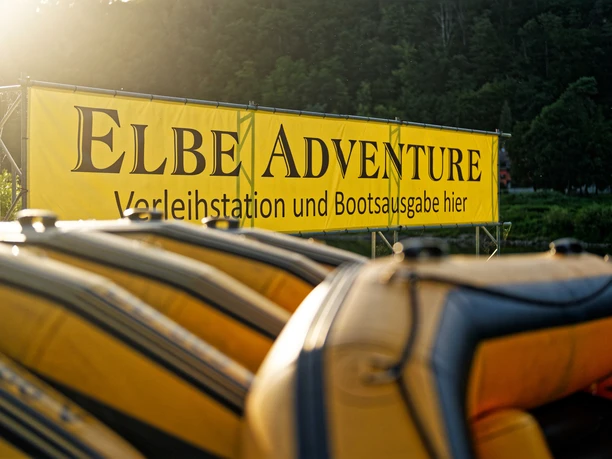 Gelbe Schlauchboote vor Banner "ELBEADVENTURE Verleihstation und Bootsausgabe hier" in grüner Landschaft.