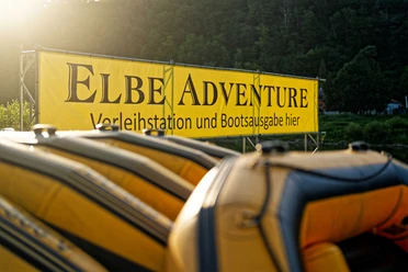 SUP_Elbe_(c)-TVSSW-Sebastian_Thiel_CC-BY-S2-TPR01026.jpg Gelbe Schlauchboote vor Banner "ELBEADVENTURE Verleihstation und Bootsausgabe hier" in grüner Landschaft.