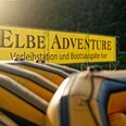 SUP_Elbe_(c)-TVSSW-Sebastian_Thiel_CC-BY-S2-TPR01026.jpg Gelbe Schlauchboote vor Banner "ELBEADVENTURE Verleihstation und Bootsausgabe hier" in grüner Landschaft.