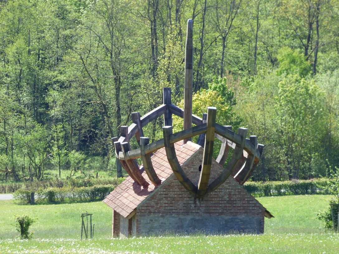 P1170785.JPG Kleines Ziegeldachhaus mit großem Holzschiffgerüst auf dem Dach, umgeben von grüner Wiese und Wald.