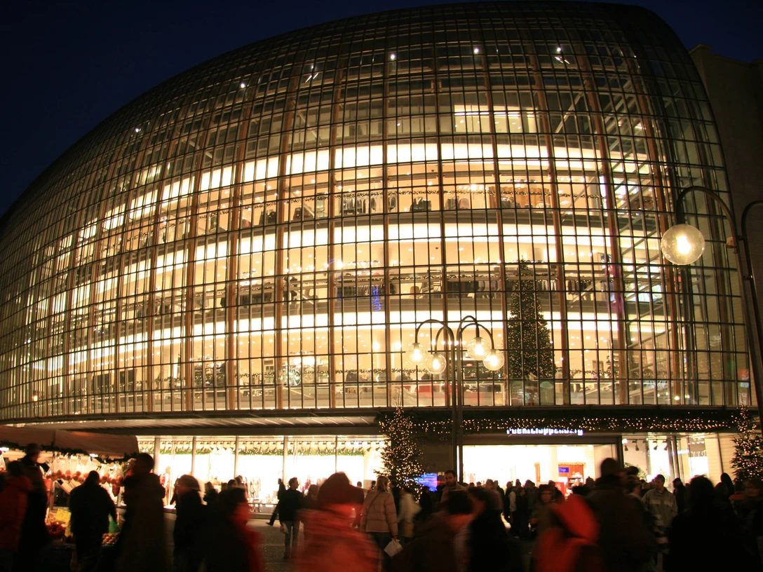 The Weltstadthaus on Schildergasse Das beleuchtete Weltstadthaus in Köln mit geschwungener Glasfassade, umgeben von Menschen.The illuminated Weltstadthaus in Cologne with its curved glass façade, surrounded by people.