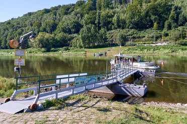 Fähre am Flussufer mit Personen, die auf das Einsteigen warten, umgeben von grünen Hügeln.