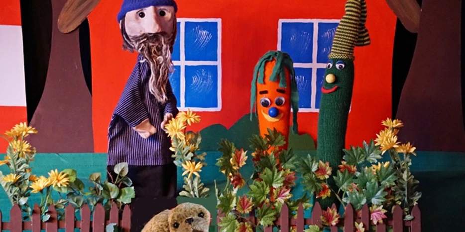 Puppen Buene Rudi Seehund 1.JPG Zwei Puppen und ein Seehund-Plüschtier stehen hinter einem Zaun mit gelben Blumen vor einer roten Wand mit zwei blauen Fenstern.