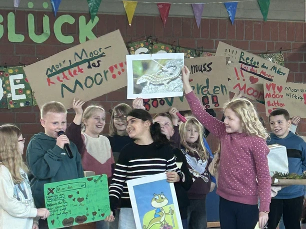 MOORex - die kleine Mooreidechse - ist das Maskottchen der Naturpark-Schulen..JPEG Kinder präsentieren fröhlich Plakate und Bilder des Maskottchens MOORex bei einer Schulfeier.