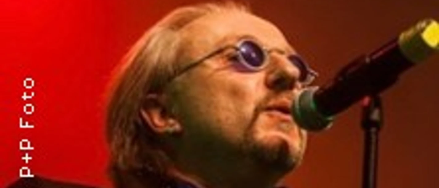 Ein Musiker mit runder Sonnenbrille singt leidenschaftlich ins Mikrofon vor rotem Licht.