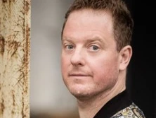 Ein Mann mit lockigem Haar trägt ein detailreich gemustertes Kleidungsstück und blickt ruhig.