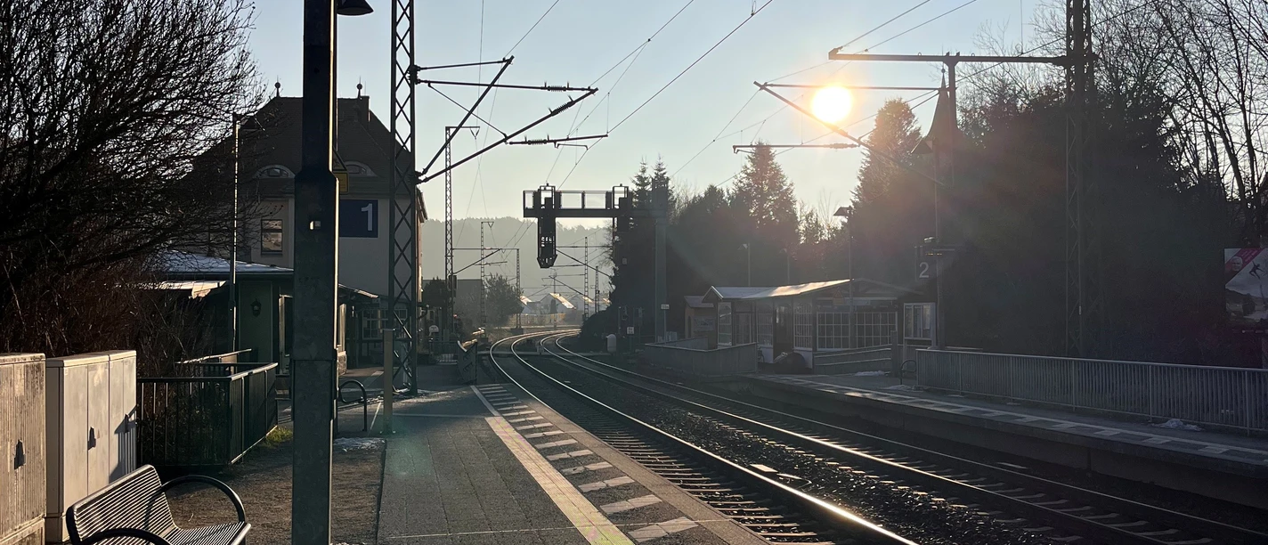 Bahnsteig mit Gleisen, Bäume und Sonne.