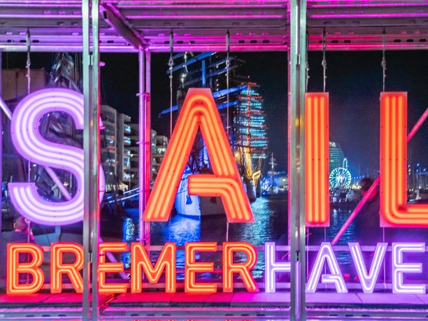 Sail Bremerhaven(c) Jens Hagens_Erlebnis Bremerhaven.jpg Leuchtendes Neon-Schild „SAIL Bremerhaven“ vor nächtlichem Hafen mit Segelschiffen und Riesenrad.