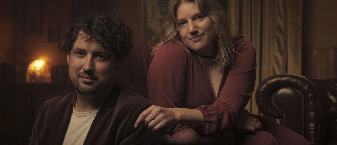 Flora.jpeg Zwei Menschen sitzen in warmem Licht auf Ledermöbeln und blicken ruhig in die Kamera.