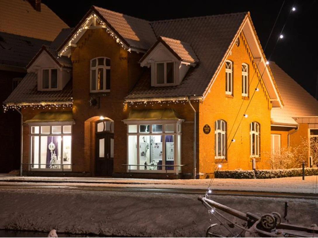 Fehntjer Forum Zweistöckiges rotes Backsteinhaus mit beleuchteten Fenstern und Dachrinnen, davor schneebedeckter Boden und ein Fahrrad im Vordergrund bei Nacht.