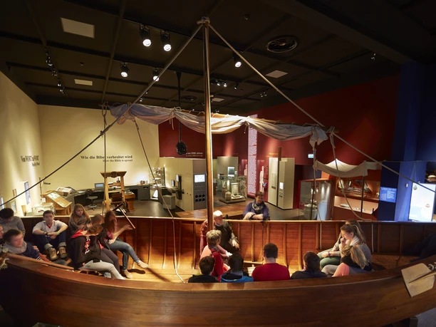 Discover the Book of Books - Ship Several people are sitting in an old wooden museum ship.