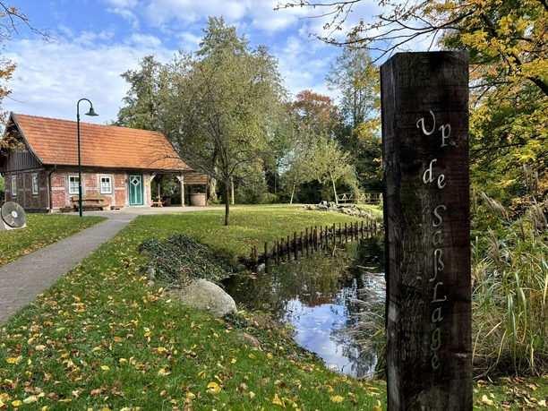Insel Saßlage, Holte-Lastrup ©Naturpark Hümmling (3) Backsteinhaus mit rotem Ziegeldach an einem Teich, umgeben von grünen Bäumen und Herbstlaub.