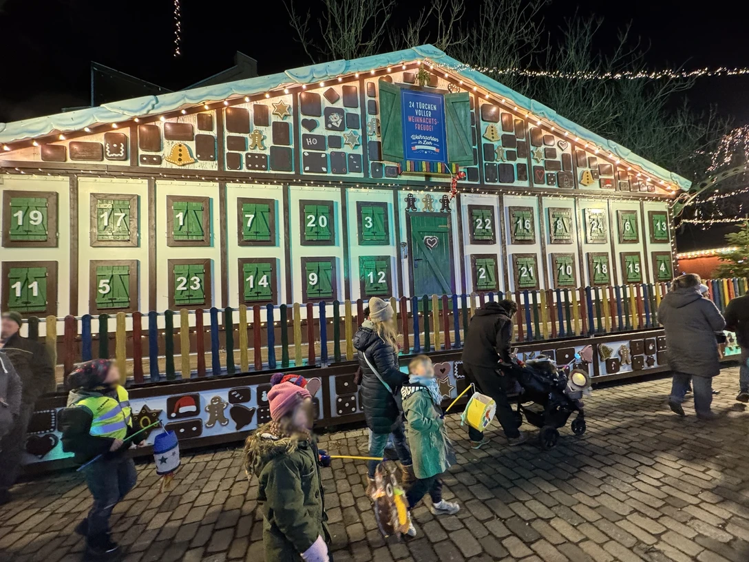 Adventskalender-weihnachtsmarkt-ernst-reuter-platz-leer-ostfriesland.jpg Leuchtendes Adventskalenderhaus mit nummerierten Fenstern, davor Familien mit Kindern und Laternen.Luminous Advent calendar house with numbered windows, in front of it families with children and lanterns.Lysende adventskalenderhus med nummererede vinduer, foran det familier med børn og lanterner.Lichtgevend adventskalenderhuis met genummerde ramen, ervoor gezinnen met kinderen en lampionnen.