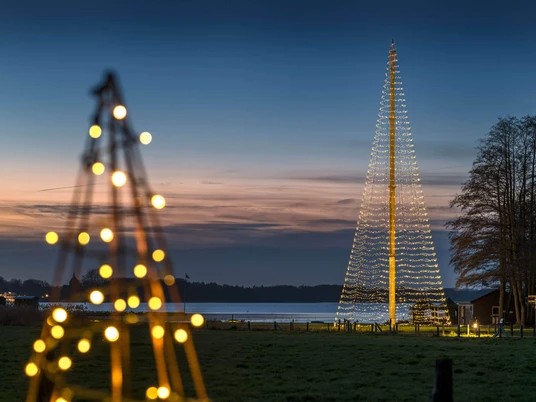 weihnachtsleuchtturm-jagdhaus-eiden.jpg Leuchtturm-Tanne aus Lichterketten am Ufer des Zwischenahner Meers bei Abenddämmerung.