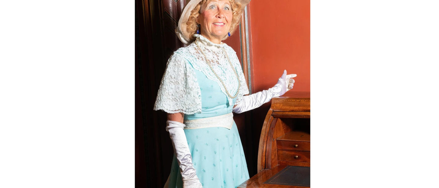 Gräfin Julia NEU Zeitreiseführung Schloss Evenburg The countess in period dress with a light blue dress and hat stands smiling next to an old writing desk