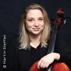 Eine Person mit Cello vor dunklem Hintergrund, entspannt sitzend und in die Kamera blickend