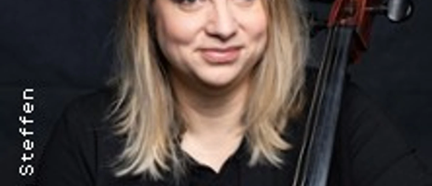 Eine Person mit Cello vor dunklem Hintergrund, entspannt sitzend und in die Kamera blickend