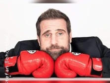 Ein Mann mit Bart stützt seinen Kopf auf zwei rote Boxhandschuhe, der Hintergrund ist weiß.