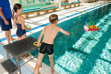 Kinder spielen fröhlich am Beckenrand eines Hallenbads und werfen Ringe auf ein buntes Wasserspiel.