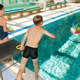 Kinderdisco Kinder spielen fröhlich am Beckenrand eines Hallenbads und werfen Ringe auf ein buntes Wasserspiel.
