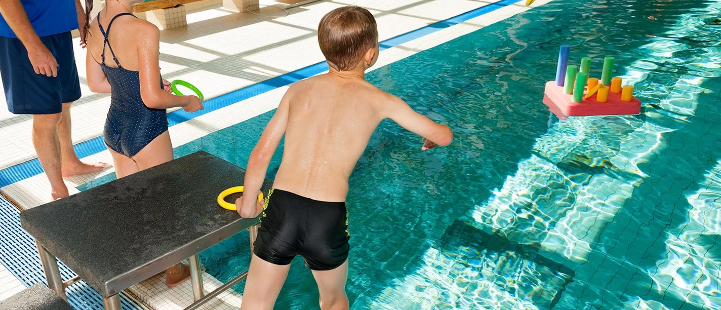 Kinder spielen fröhlich am Beckenrand eines Hallenbads und werfen Ringe auf ein buntes Wasserspiel.