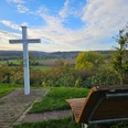 Weiße Kreuzsäule an einem Weg mit Holzbank, Blick auf Felder und Dorf in hügeliger Landschaft.