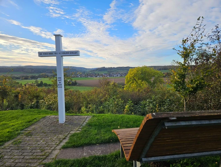 Emder Höhe Weiße Kreuzsäule an einem Weg mit Holzbank, Blick auf Felder und Dorf in hügeliger Landschaft.