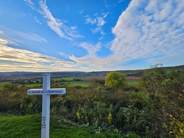 Weißes Kreuz mit Inschrift vor weitem Blick über Felder, Wiesen und Wälder bei Bad Driburg-Alhausen.