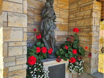 Erpentrup Bildstock Bronzestatue einer Frau mit Kind vor Sandsteinwand, umrahmt von roten und weißen Blumenkästen.