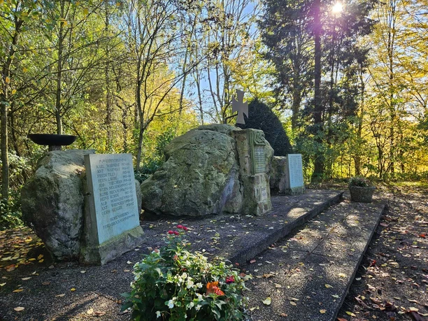 Steindenkmal mit Namenstafeln für Gefallene, umgeben von Bäumen im Sonnenlicht und Blumen im Vordergrund.