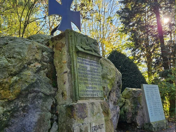 Gedenkstein aus Naturstein mit schwarzem Kreuz und Namenstafeln, umgeben von Bäumen im Sonnenlicht.